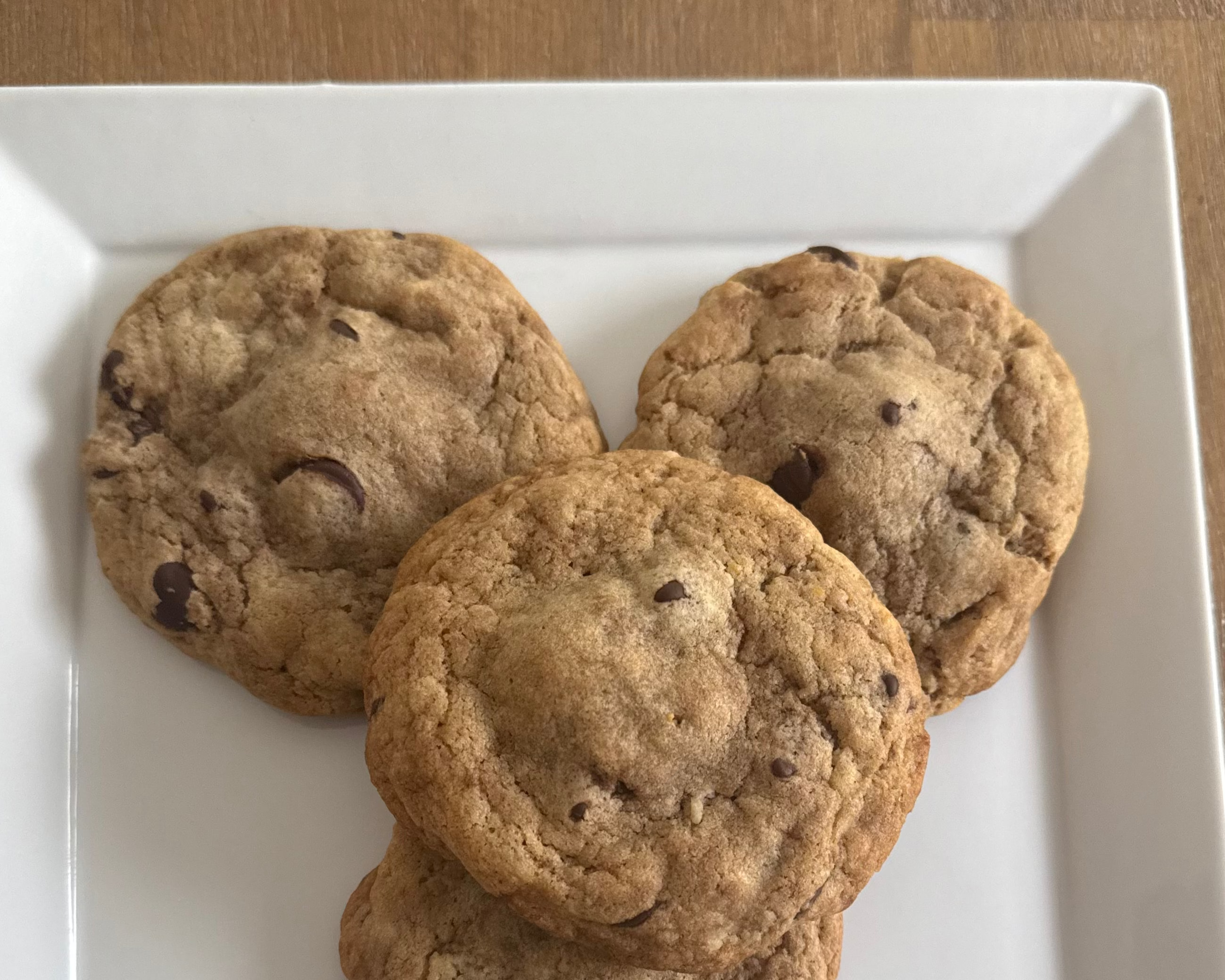 Four chocolate chip cookies on a white plate with a wooden surface background